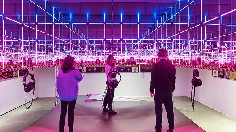 Drei Besucher stehen in einem Raum des Vitra-Designmuseums inmitten pinker und blauer Neonröhren, die ein violettes Licht machen. Kopfhörer hängen von der Decke.