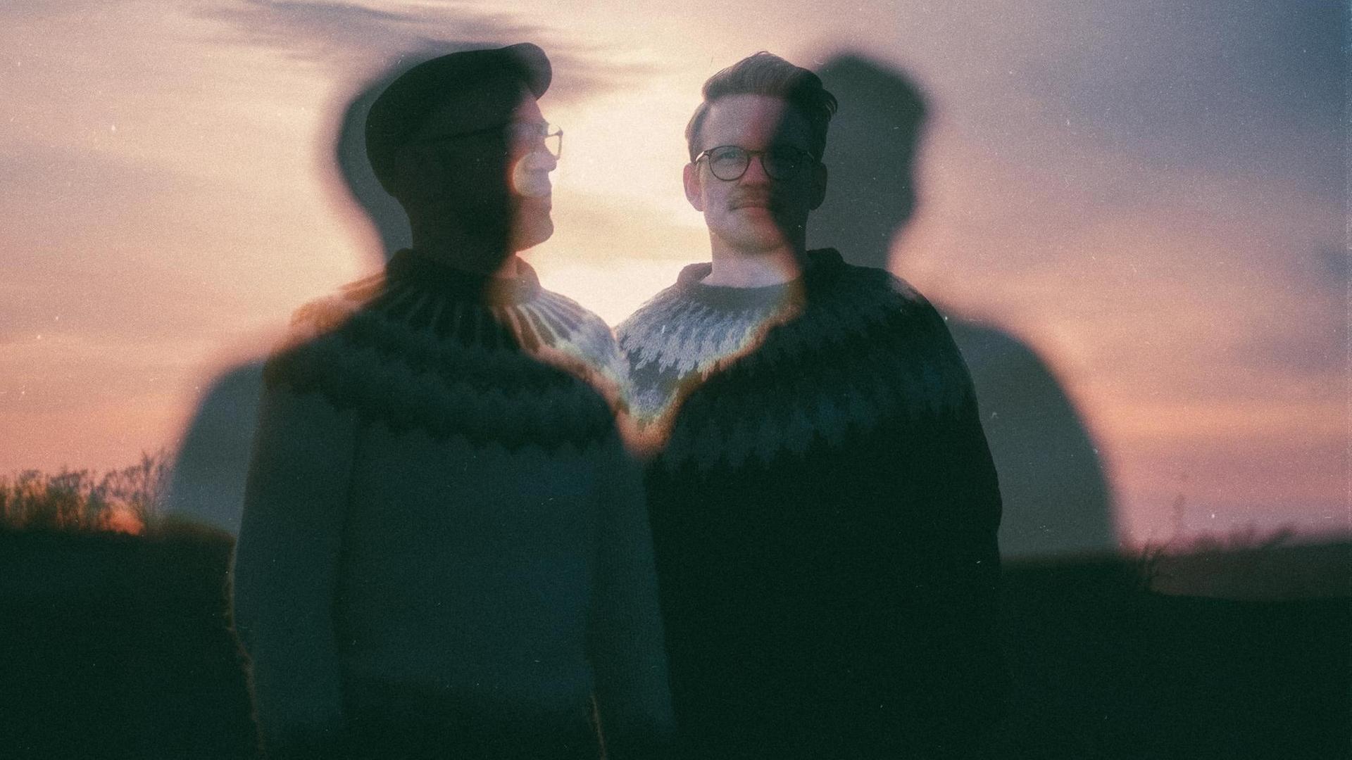 Auf dem Bild sind die verschwommenen Umrisse zweier Männer zu sehen, die im Dämmerlicht draußen stehen. Sie tragen Island-Pullover. Auf dem Bild sind die verschwommenen Umrisse zweier Männer zu sehen, die im Dämmerlicht draußen stehen. Sie tragen Island-Pullover.