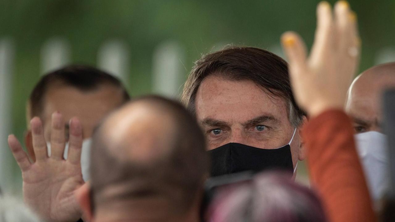 Großaufnahme von Präsident Bolsonaro, der einen schwarzen Mund-Nasenschutz trägt. Sein Gesicht wird halb verdeckt von Anhängern, die ihm zuwinken. Großaufnahme von Präsident Bolsonaro, der einen schwarzen Mund-Nasenschutz trägt. Sein Gesicht wird halb verdeckt von Anhängern, die ihm zuwinken.