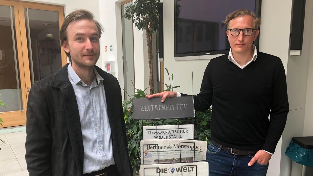 Zwei Männer stehen in einem Büro, zwischen ihnen ist ein Zeitungsständer. Zwei Männer stehen in einem Büro, zwischen ihnen ist ein Zeitungsständer.