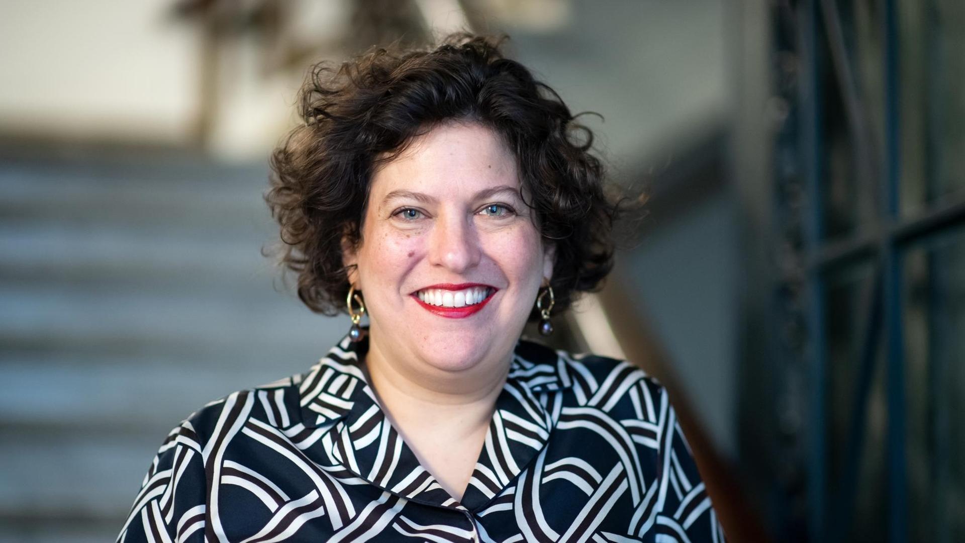 Ruth Ur, mit dunklen Locken und blauen Augen und dunkler Bluse mit Muster, steht im Treppenhaus einer Synagoge in Berlin-Charlottenburg. Ruth Ur, mit dunklen Locken und blauen Augen und dunkler Bluse mit Muster, steht im Treppenhaus einer Synagoge in Berlin-Charlottenburg.