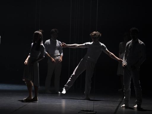 Symbolbild: Tänzerinnen und Tänzer bei einer Probe, in weißer Kleidung im Zwielicht auf einer dunklen Bühne. 