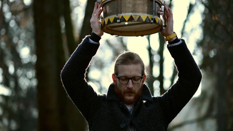 Ein juner Mann mit Brille und Vollbart steht in einem Wald und hebt mit beiden Armen eine Trommel über seinen Kopf, während er nach unten schaut. Ein juner Mann mit Brille und Vollbart steht in einem Wald und hebt mit beiden Armen eine Trommel über seinen Kopf, während er nach unten schaut.