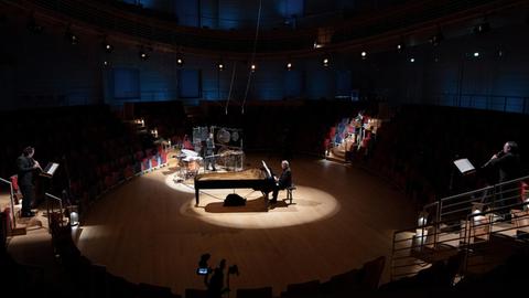 Ein Mann sitzt an einem Flügel in der Mitte des Pierre-Boulez-Saals in Berlin, auf den Treppen des Saals stehen Musiker mit Streich- und Blasinstrumenten, im Vordergrund eine Kamera.