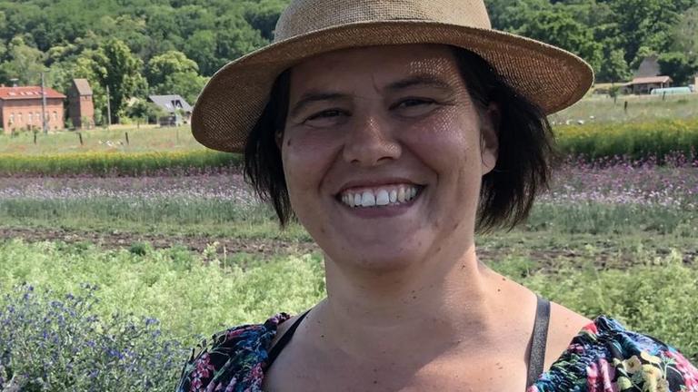 Wildblumenzrüchterin Nina Keller steht mit einem Strohhut auf dem Kopf vor einem ihrer Wildblumenfelder, im Hintergrund ein Dorf im Oderbruch in Brandenburg. Wildblumenzrüchterin Nina Keller steht mit einem Strohhut auf dem Kopf vor einem ihrer Wildblumenfelder, im Hintergrund ein Dorf im Oderbruch in Brandenburg.