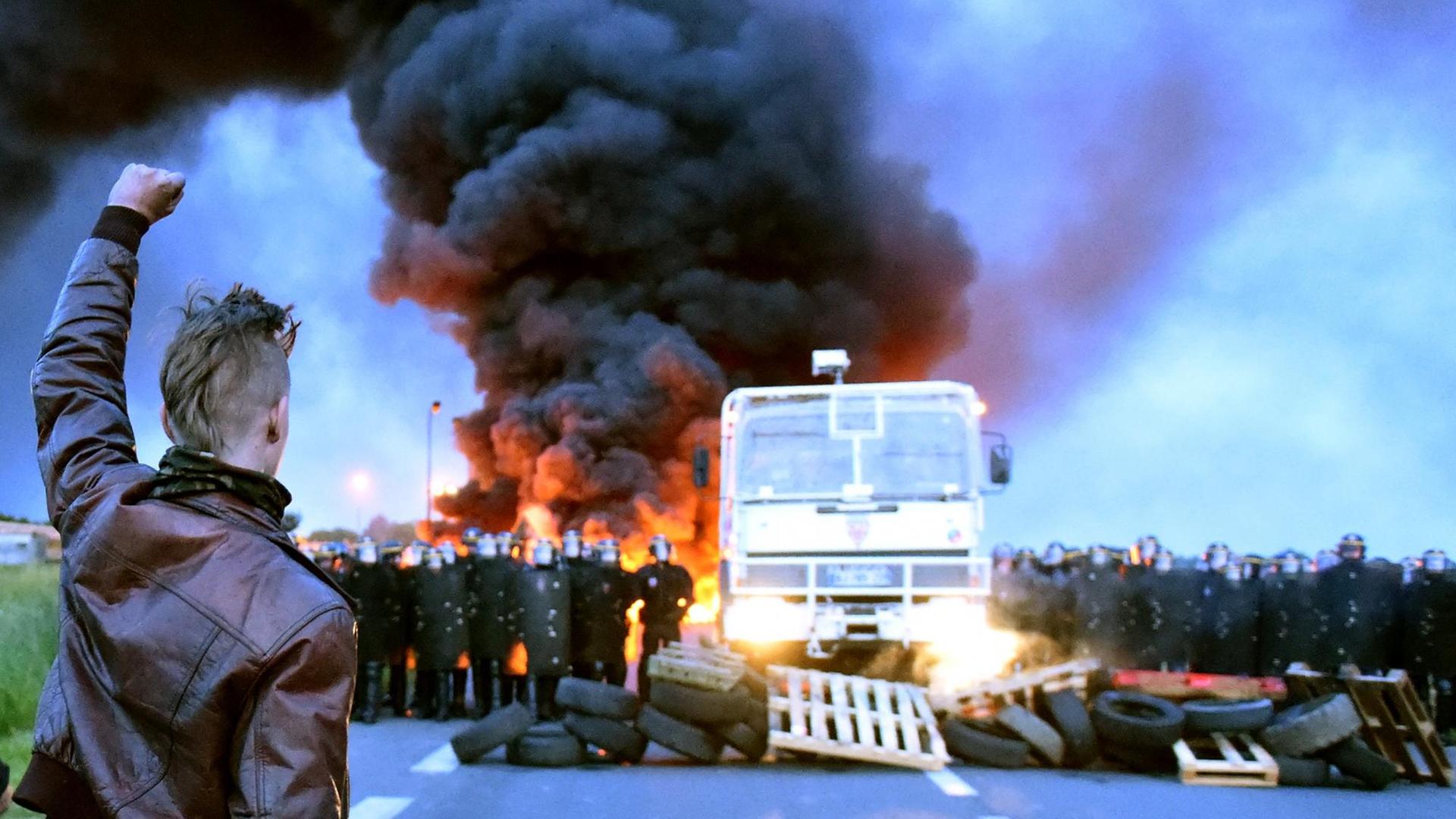 Ein Demonstrant hält die Hand in die Luft, im Hintergrund eine Blockade aus Reifen auf einer Straße, Polizisten sperren die Straße, dahinter ist ein Feuer zu sehen. Ein Demonstrant hält die Hand in die Luft, im Hintergrund eine Blockade aus Reifen auf einer Straße, Polizisten sperren die Straße, dahinter ist ein Feuer zu sehen.