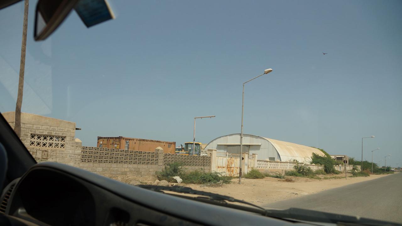 Die Baufirma in Eritrea, bei der Zenagabriel gearbeitet hat: Auf einem Auto heraus sieht man neben einer Straße eine Lagerhalle und mehrere andere Gebäude. Die Baufirma in Eritrea, bei der Zenagabriel gearbeitet hat: Auf einem Auto heraus sieht man neben einer Straße eine Lagerhalle und mehrere andere Gebäude.