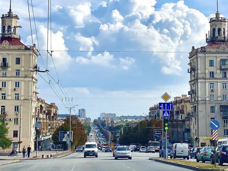 In der Innenstadt von Saporischschja warten Autos an einer Ampelkreuzung. Links ist eine Oberleitung für eine Straßenbahn. Wolken sind am Himmel.