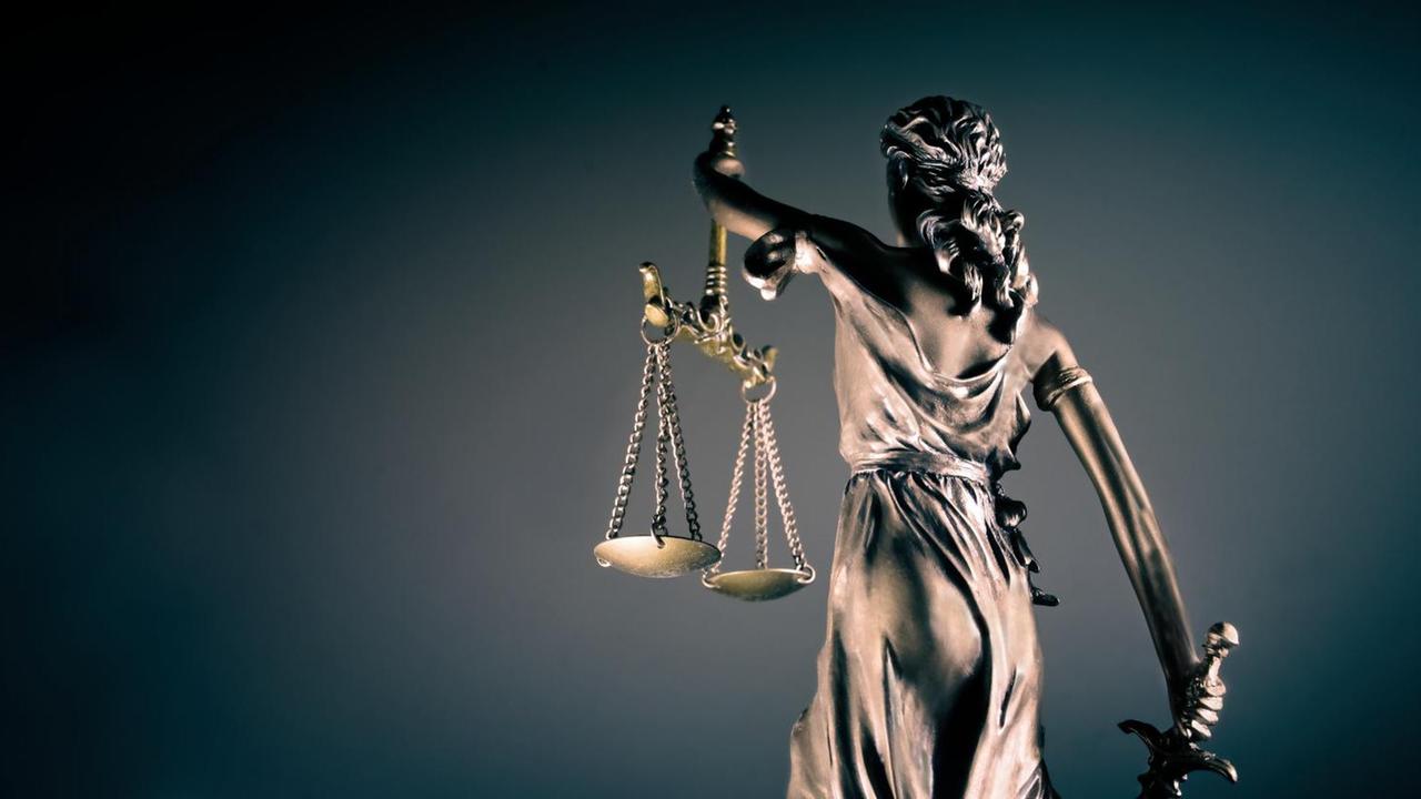 Vor einem dunklen Hintergrund steht dem Betrachter mit dem Rücken zugewandt die Figur der Justitia, die in der rechten Hand ein Schwert und in der linken Hand zwei Waageschalen trägt. Vor einem dunklen Hintergrund steht dem Betrachter mit dem Rücken zugewandt die Figur der Justitia, die in der rechten Hand ein Schwert und in der linken Hand zwei Waageschalen trägt.