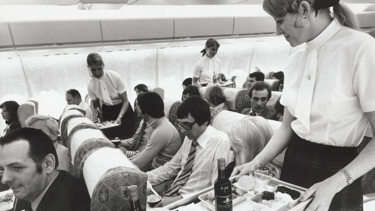 Blick in die Economy-Klasse in den 70er-Jahren. Drei Stewardessen bewirten die Passagiere in einem Flugzeug. 
