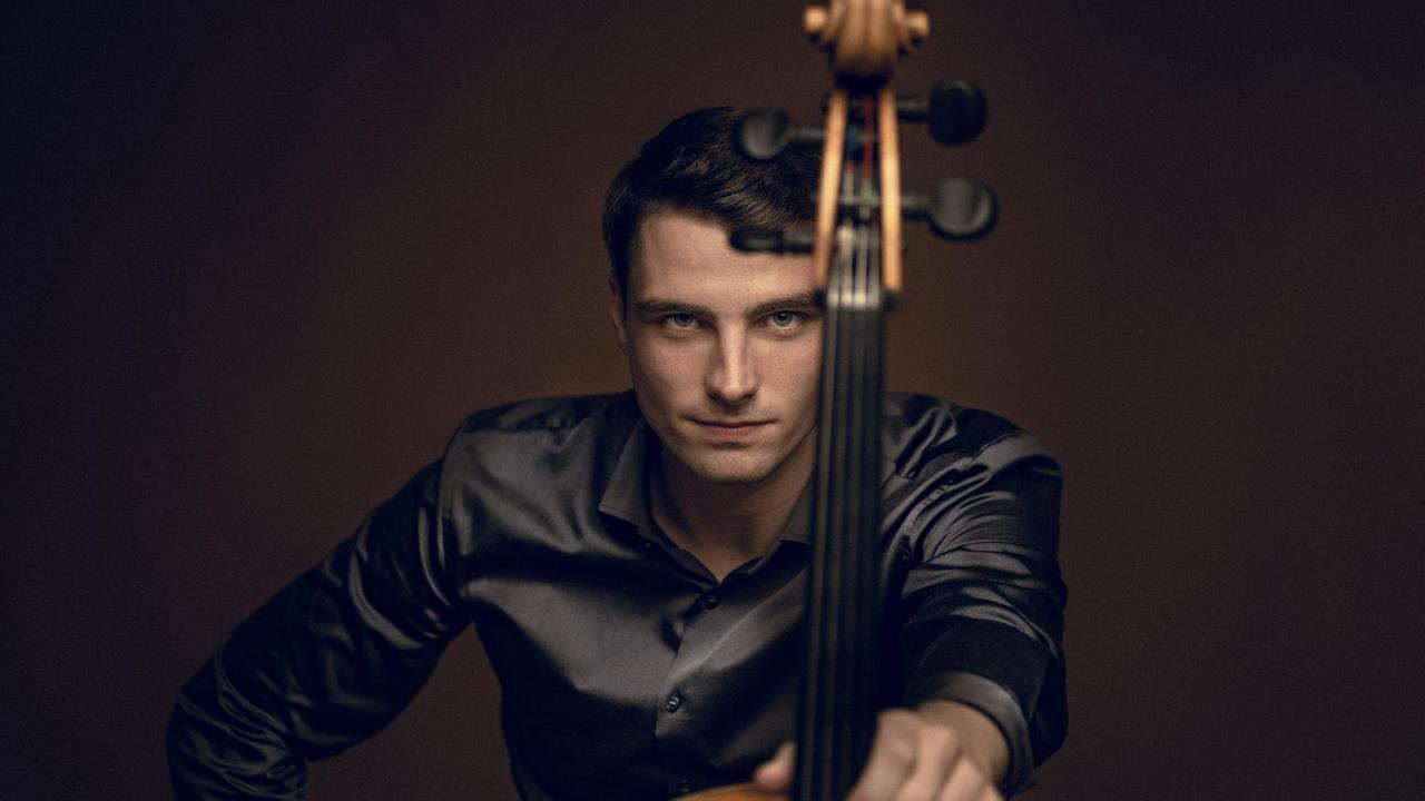 Cellist Friedrich Thiele blickt direkt in die Kamera, während er sein Cello mit ausgestrecktem Arm vor sich hält.