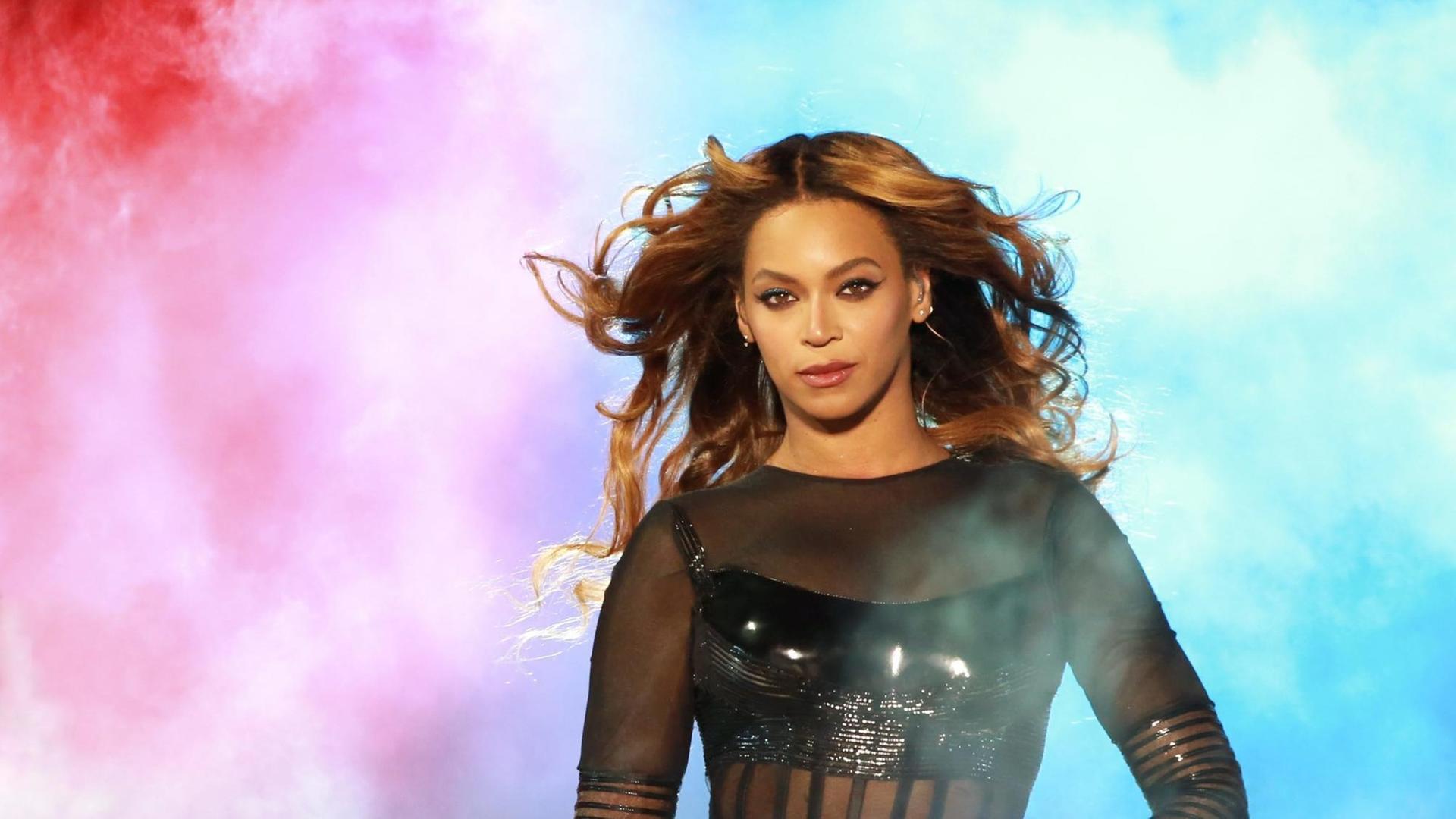 Beyoncé läuft bei einem Auftritt durch rot und blau angeleuchteten Nebel. Sie trägt einen schwarzen, teilweise durchsichtigen Body. Beyoncé läuft bei einem Auftritt durch rot und blau angeleuchteten Nebel. Sie trägt einen schwarzen, teilweise durchsichtigen Body.