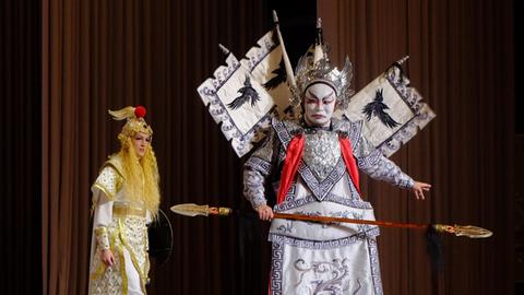 Auf der Bühne rechts im Vordergrund steht ein chinesischer Darsteller mit weiß geschminktem Gesicht in einem üppigen silbernen Kostüm mit Lanze in der Hand, im Hintergrund links eine Darstellerin im goldenen Kleid mit langen blonden Haaren