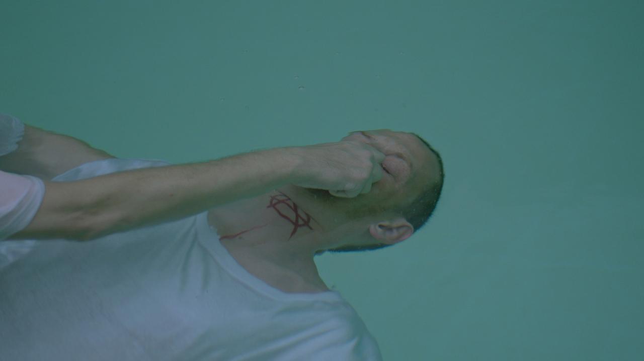 Im Still aus "Jesus Egon Christus" wird ein Mann im weißen T-Shirt und mit einem roten Anarchiesymbol auf dem Hals unter Wasser getaucht.