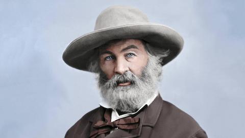Ein coloriertes Porträt des Dichters Walt Whitman. Er sitzt mit einem Hut mit breiter Krempe auf einen Stuhl und schaut in die Kamera. Seine Hände sind in den Manteltaschen.