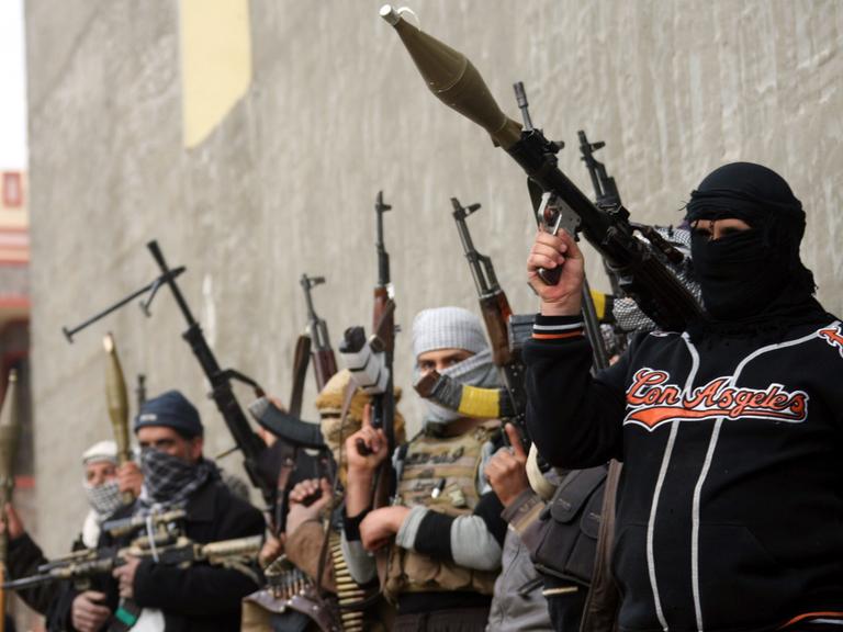 Vermummte, mit Raketenwerfern und Maschinengewehren bewaffnete Männer stehen vor einer Mauer.
