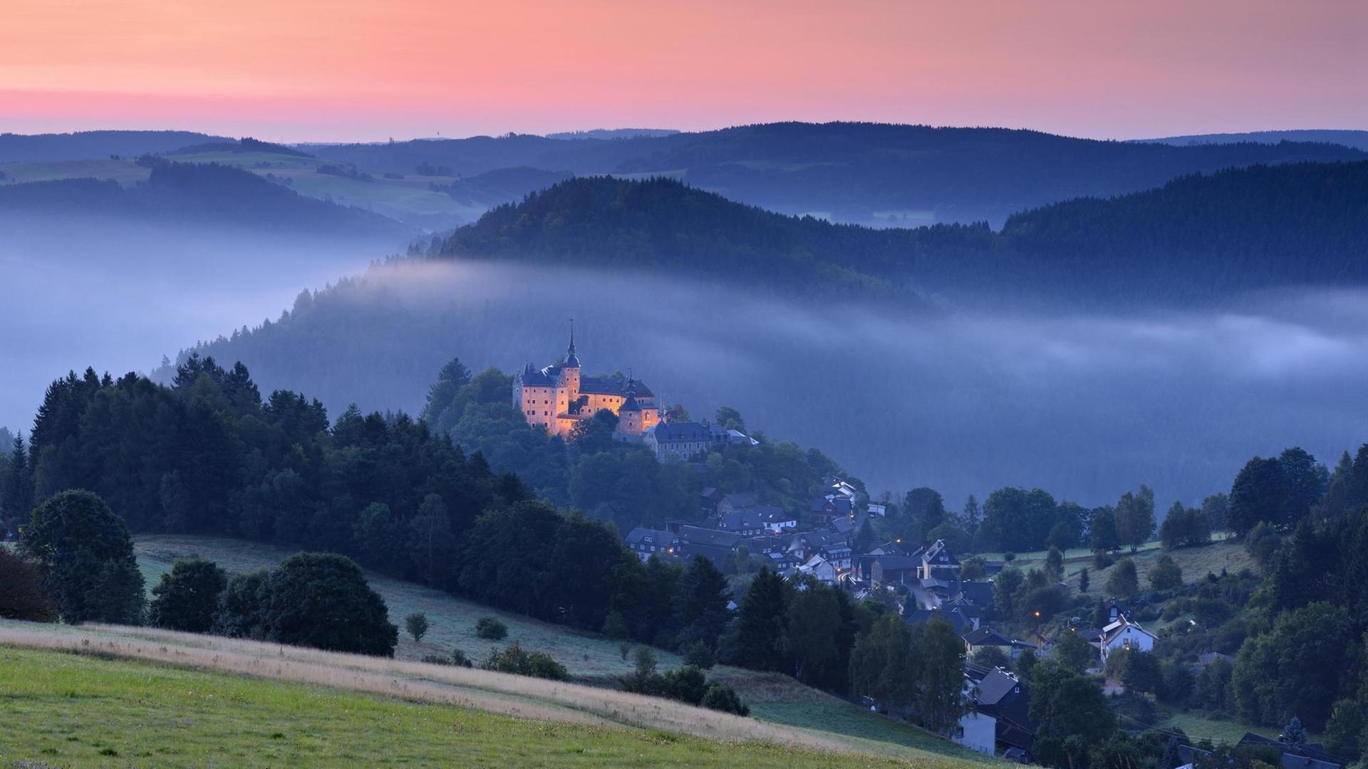 Die Burg Lauenstein liegt von Morgennebel umgeben über dem Dorf, darüber der Himmel glüht in den Farben des Sonnenaufgangs.