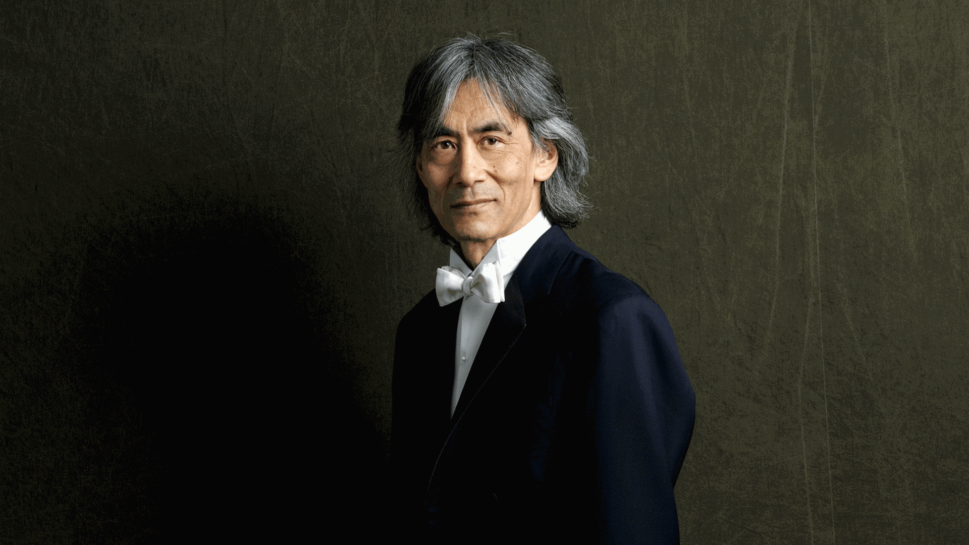 Kent Nagano steht vor einer getäfelten Wand und blickt in Abendgarderobe in die Kamera. Kent Nagano steht vor einer getäfelten Wand und blickt in Abendgarderobe in die Kamera.