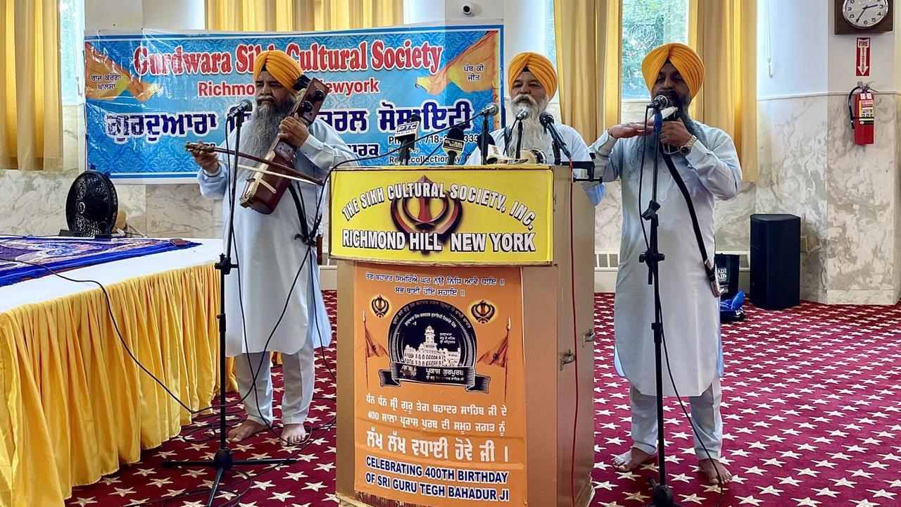 Drei Musiker in weißen Gewändern und mit gelben Turbanen treten in der Gebetshalle eines New Yorker Sikh-Tempels auf. Sie spielen auf Trommeln und einem Streichinstrument. Das Plakat auf einem Stehpult weist auf die Feier zum 400. Geburtstag eines Gurus hin.