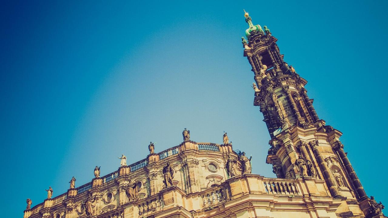 Blick von Unten zum Kirchenschiff und Turmspitze der Dresdner Hofkirche mit ihren vielen Figuren auf dem Dach und der gelb-grauen Sandsteinfassade. Blick von Unten zum Kirchenschiff und Turmspitze der Dresdner Hofkirche mit ihren vielen Figuren auf dem Dach und der gelb-grauen Sandsteinfassade.
