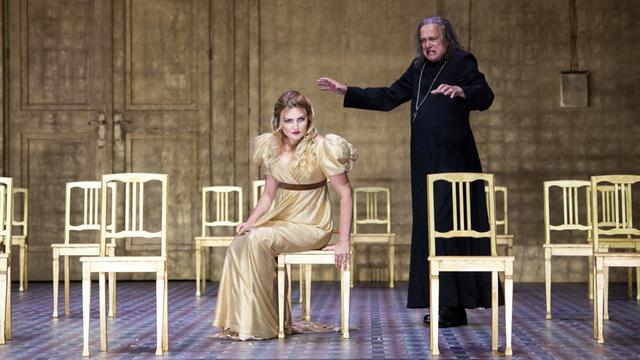 Eine Frau mit blonden langen Haaren sitzt auf einem von mehreren goldenen Stühlen, dahinter steht ein schwarzgekleideter Mann und hebt singend die Arme.