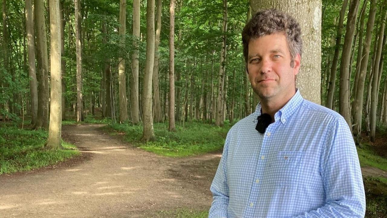 Sebastian Graf von Platen steht im Wald. Er trägt eine olivgrüne Arbeitshose und ein hellblau-weiß kariertes Hemd. Sebastian Graf von Platen steht im Wald. Er trägt eine olivgrüne Arbeitshose und ein hellblau-weiß kariertes Hemd.
