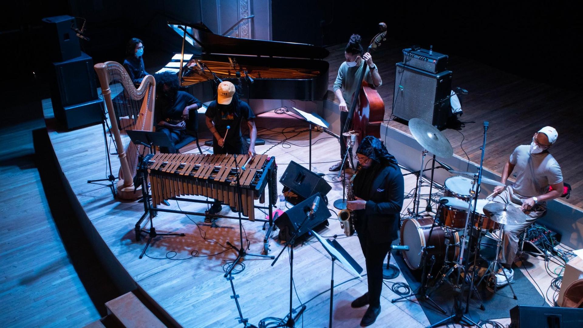 Aus der Vogelperspektive sind sechs Musiker mit einem Mund-Nasen-Schutz zu sehen, die auf einer blau ausgeleuchteten Bühne spielen.