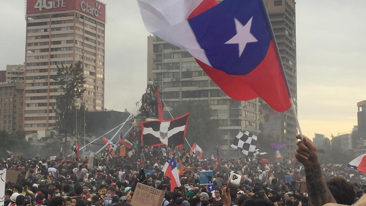 Ein Platz in Santiago de Chile, voll mit Menschen, die die chilenische Fahne schwenken, eingerahmt von Hochhäusern.