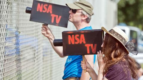 Mit zwei Pappkameras mit der Aufschrift "NSA TV" schauen zwei Teilnehmer einer Protestkundgebung vor dem "Dagger Complex" des US-Geheimdiensts NSA in Griesheim bei Darmstadt (Hessen) am 26.07.2014 durch den Sicherheitszaun der Anlage.