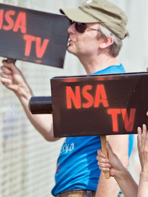 Mit zwei Pappkameras mit der Aufschrift "NSA TV" schauen zwei Teilnehmer einer Protestkundgebung vor dem "Dagger Complex" des US-Geheimdiensts NSA in Griesheim bei Darmstadt (Hessen) am 26.07.2014 durch den Sicherheitszaun der Anlage.