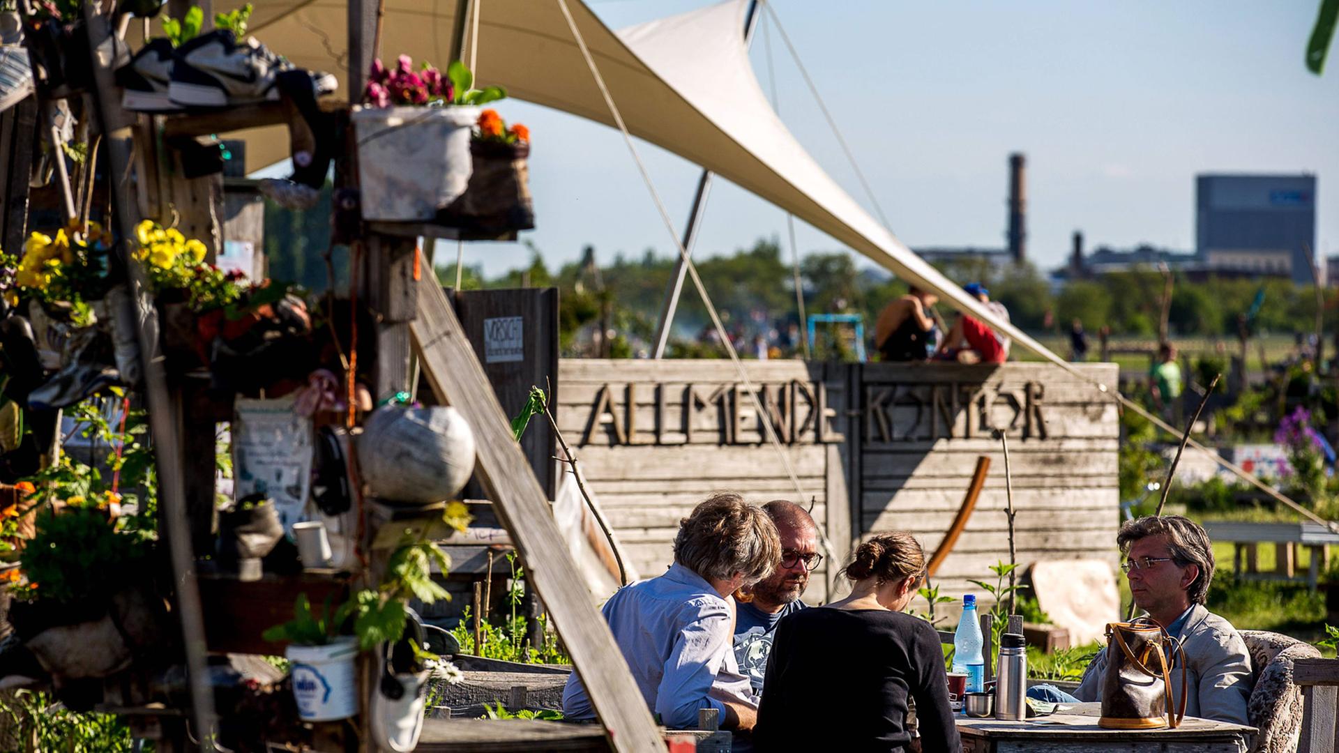 Eine Gruppe Menschen sitzen von Pflanzen und Holzbauten umgeben in einem Garten auf dem Feld des ehemaligen Flughafens Tempelhof in Berlin. Eine Gruppe Menschen sitzen von Pflanzen und Holzbauten umgeben in einem Garten auf dem Feld des ehemaligen Flughafens Tempelhof in Berlin.