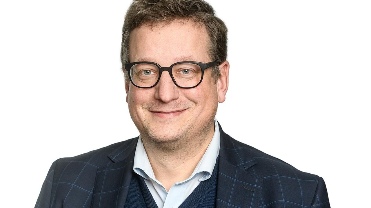 Ein bebrillter Mann in Hemd und Sakko steht vor einem weißen Hintergrund und lächelt in die Kamera. Ein bebrillter Mann in Hemd und Sakko steht vor einem weißen Hintergrund und lächelt in die Kamera.