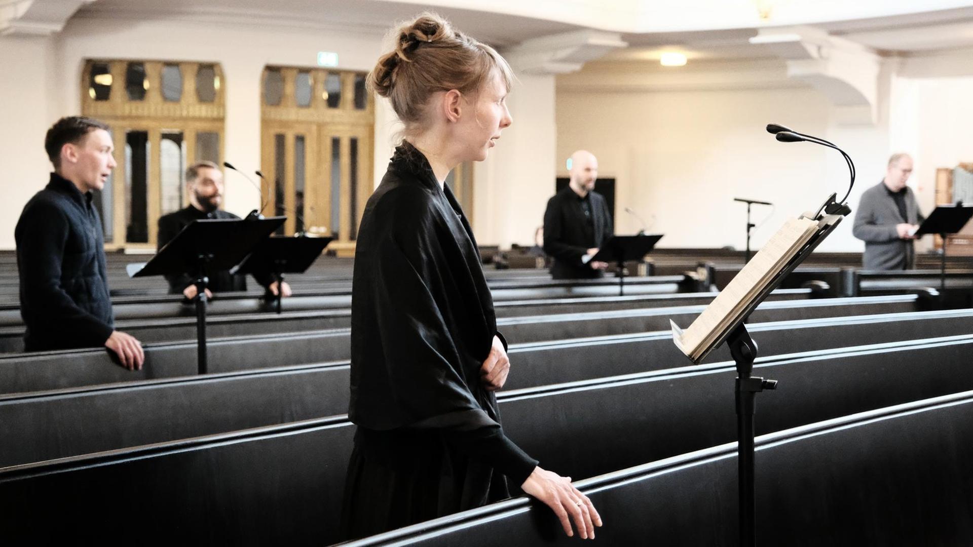 Im Vordergrund ist eine schwarz gekleidete Sängerin zu sehen, die in einer dunklen Kirchenbank steht. Im Vordergrund ist eine schwarz gekleidete Sängerin zu sehen, die in einer dunklen Kirchenbank steht.
