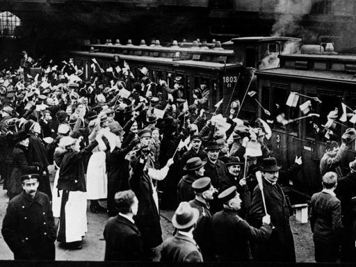 Ein Schwarzweiß-Foto zeigt eine große Menschenmenge am Gleis in einer großen Bahnhofshalle ,zum Großteil Fahnen schwenkend vor einem Zug aus zwei Waggons. Links sind zahlreiche Frauen in weißen Röcken, vermutlich einer Tracht zu sehen die dem Zug entgegen winken. Auf der rechten Bildhälfte bfinden sich zahlreiche Männer in Anzug und mit Schirmmützen Ein Sonderzug für Stimmberechtigte bei der Volksabstimmung Schlesien am 20. Maäz 1921 wartet im Schlesischen Bahnhof Berlin