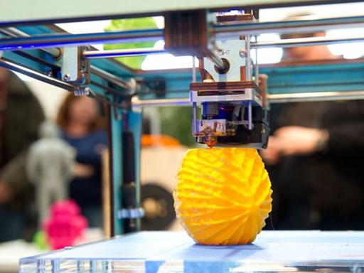 Ein 3D-Drucker druckt ein gelbes kugelförmiges Gebilde aus.