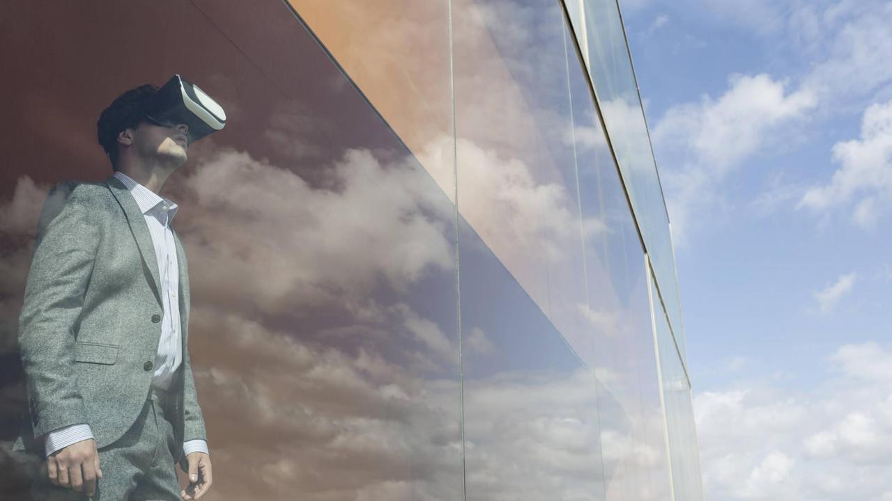 Ein Mann im Anzug und einer VR-Brille steht an einem Fenster eines Hochhauses, in dem sich der Himmel spiegelt. Ein Mann im Anzug und einer VR-Brille steht an einem Fenster eines Hochhauses, in dem sich der Himmel spiegelt.