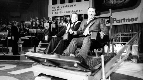 Wilfried Dietrich und Rudolf Mang sitzen auf der Bühne angeschnallt in Autositzen.