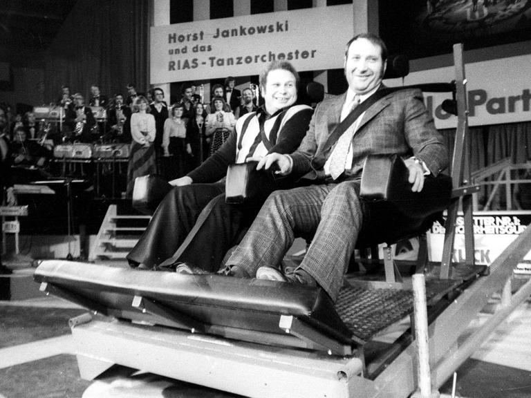 Wilfried Dietrich und Rudolf Mang sitzen auf der Bühne angeschnallt in Autositzen.
