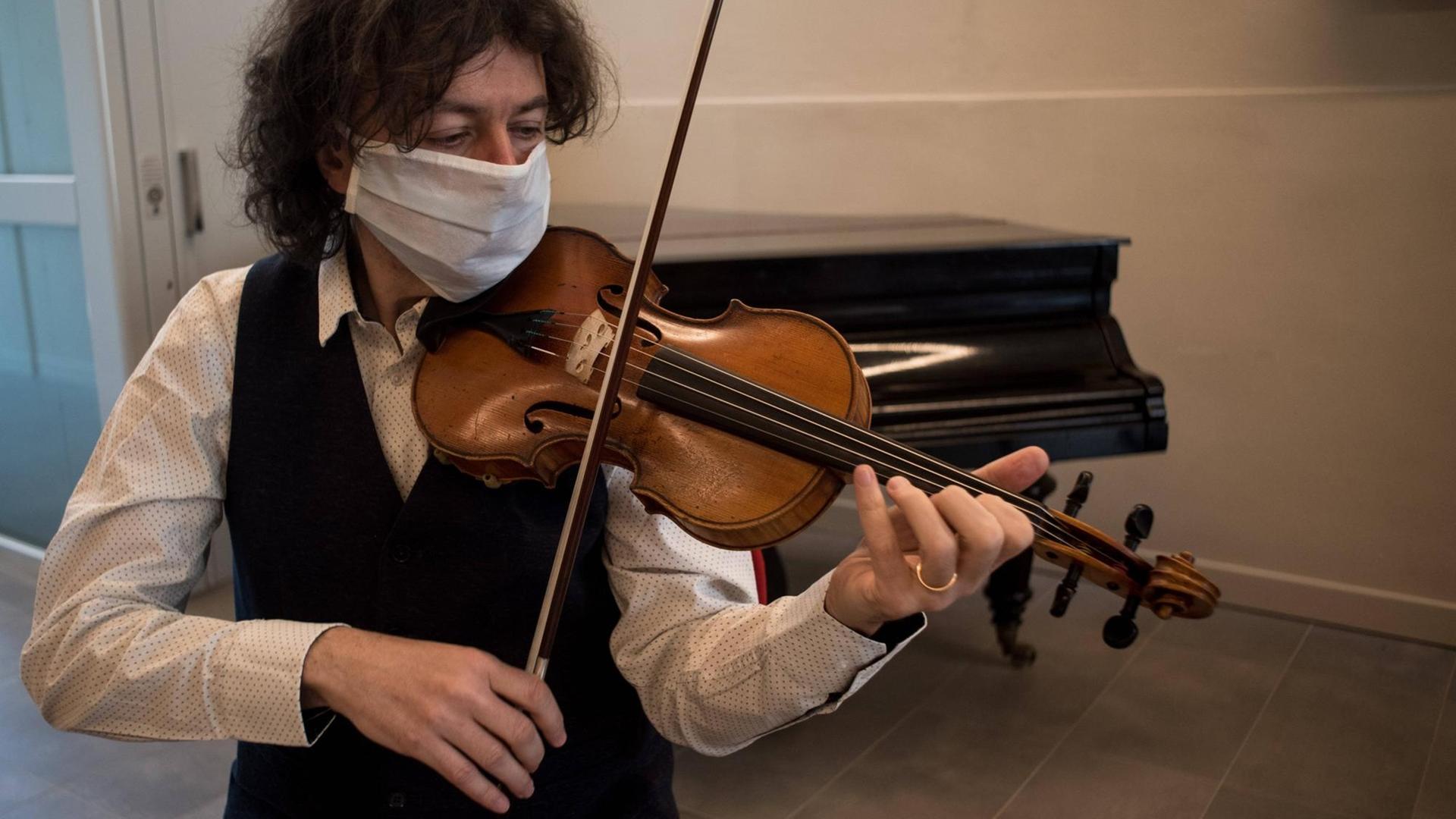 Ein Geiger sitzt mit Maske in einem kargen Unterrichtsraum und übt vor einem geschlossenen Flügel. Ein Geiger sitzt mit Maske in einem kargen Unterrichtsraum und übt vor einem geschlossenen Flügel.