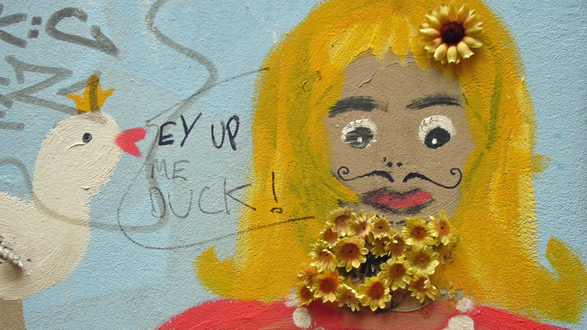 Blick auf ein Grafitti einer Frau, mit künstlichen Blumen wurde ein Bart angeklebt