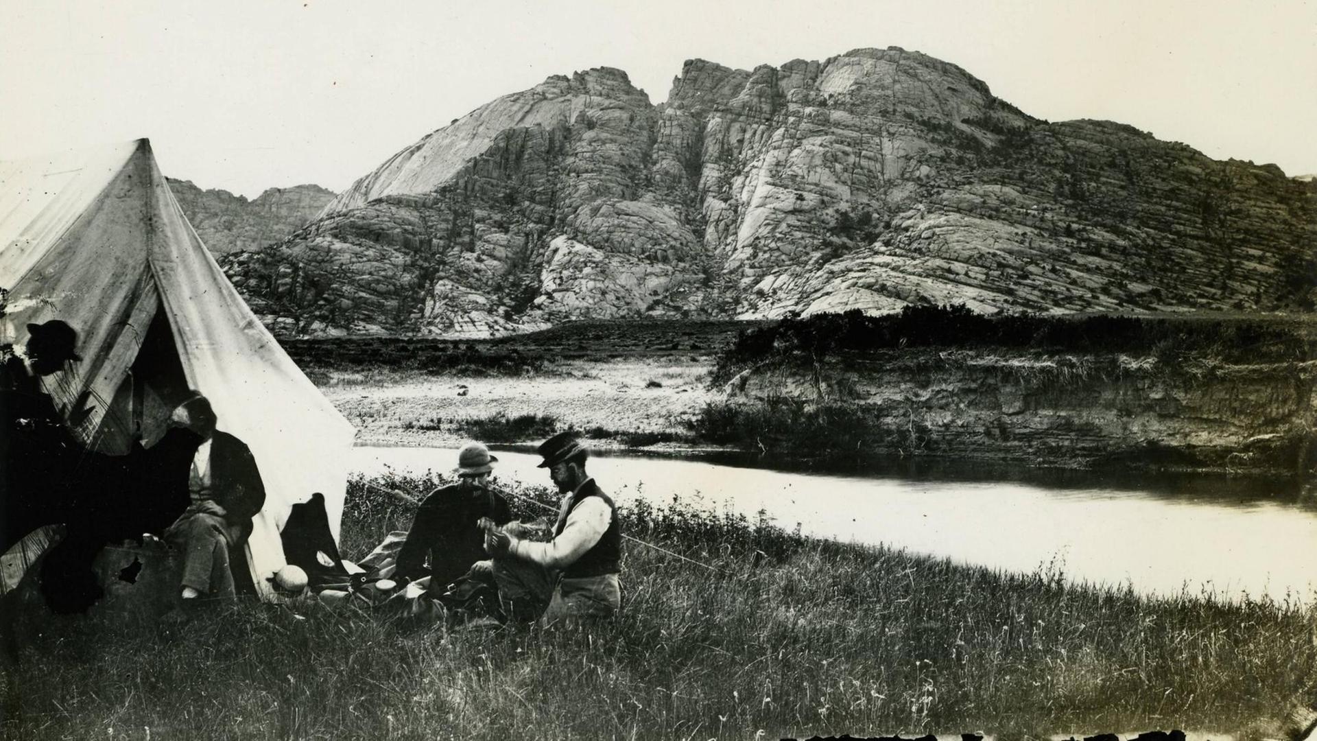 Ein vergilbtes Schwarzweiß-Foto zeigt drei Männer vor einem Zelt am Ufer eines breiten Flusses. Den Hintergrund des Bildes füllt ein, in der Mitte gespaltener Felsrücken aus