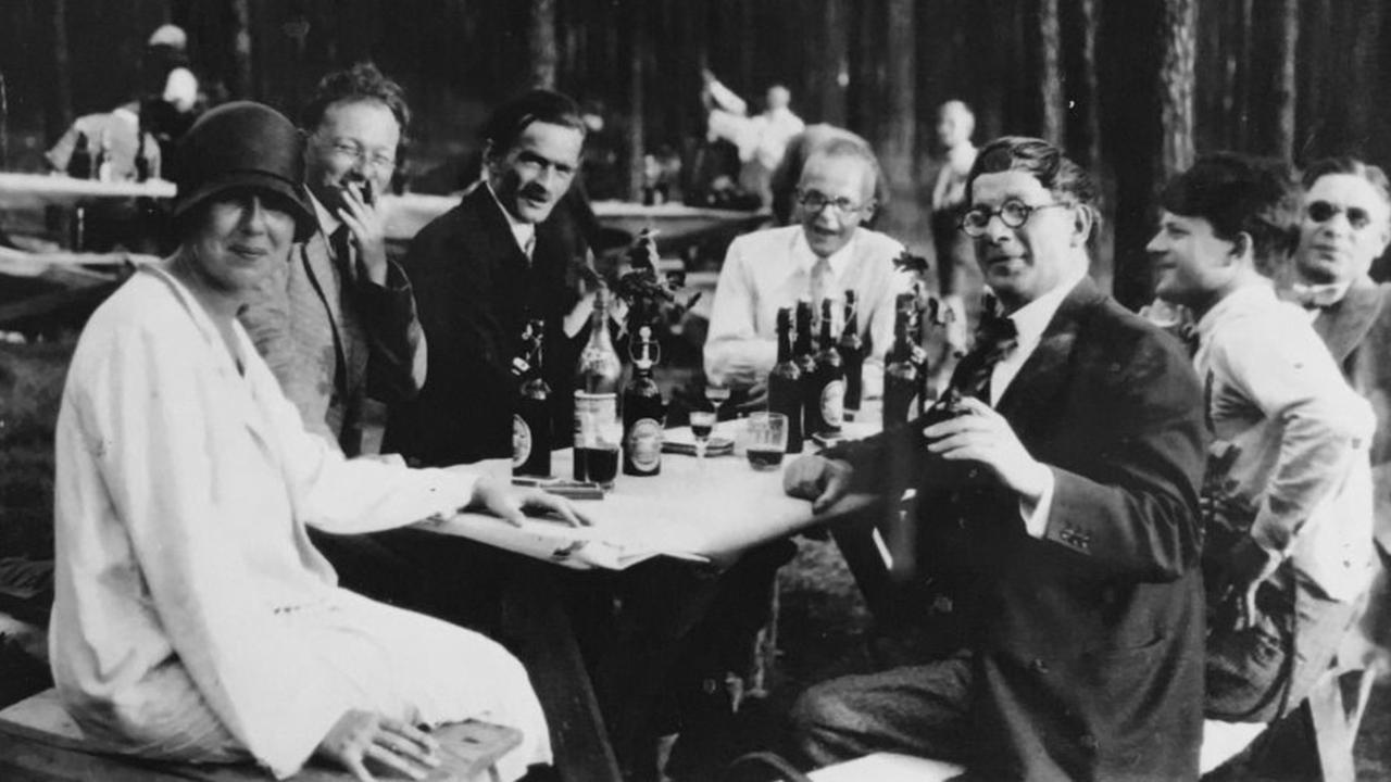 Schwarz-weiß Foto von 6 Männern und einer Frau. Sie sitzen um einem Tisch im Freien, auf dem Bierflaschen stehen.