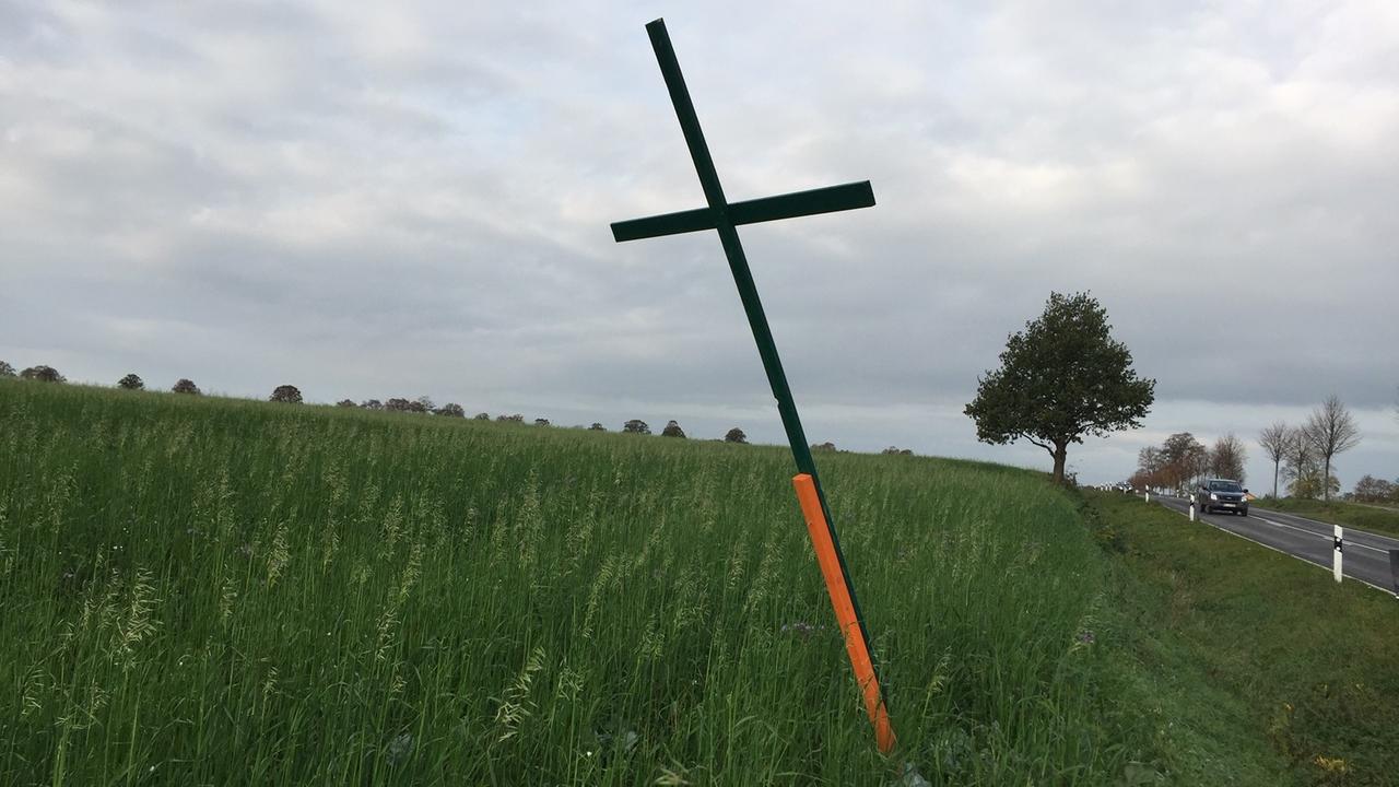Ein grünes Holzkreuz steht auf einer Wiese neben einer Straße. Ein grünes Holzkreuz steht auf einer Wiese neben einer Straße.