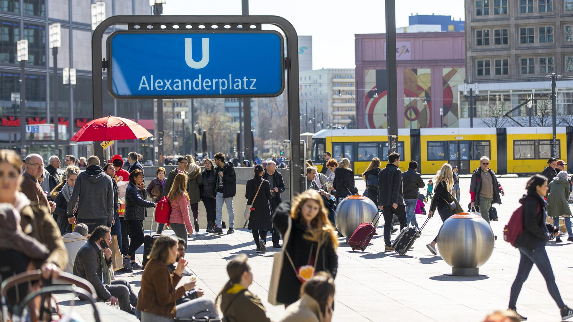 Auf einem belebten Platz mit einem Abgang zur U-Bahnhaltestelle Alexanderplatz sind viele Menschen unterwegs.