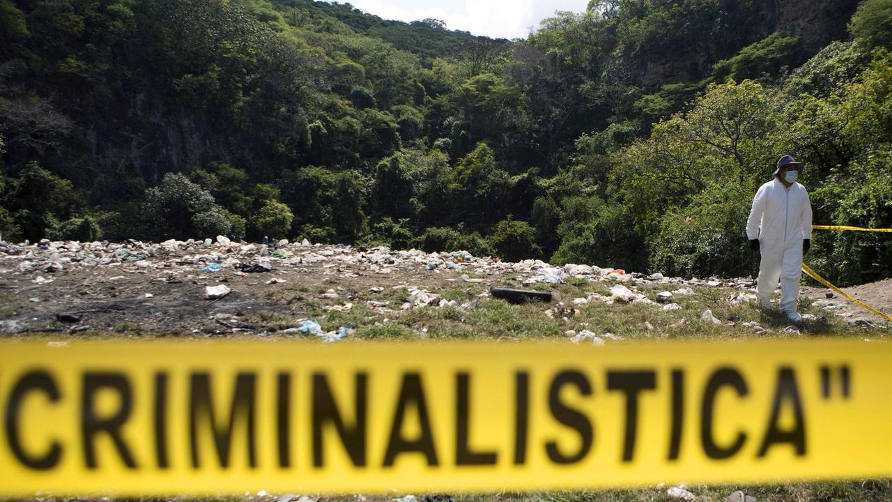 Hinter einer gelben Absperrung mit der schwarzen Aufschrift "Criminalistica" sieht man eine Müllkippe mit einem in weiße Schutzkleidung gehüllten Forensiker, im Hintergrund ein bewaldeter Hügel. Hinter einer gelben Absperrung mit der schwarzen Aufschrift "Criminalistica" sieht man eine Müllkippe mit einem in weiße Schutzkleidung gehüllten Forensiker, im Hintergrund ein bewaldeter Hügel.
