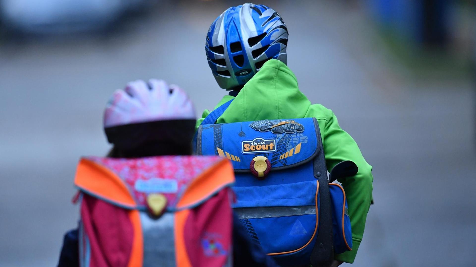 Zwei Kinder mit Schulranzen und Fahrradhelmen sind von hinten auf ihren Fahrrädern auf dem Schulweg zu sehen. Zwei Kinder mit Schulranzen und Fahrradhelmen sind von hinten auf ihren Fahrrädern auf dem Schulweg zu sehen.