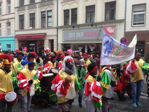 Eine grün-gelb gekleidete Karnevalsgruppe läuft auf einer Straße in einem Umzug mit, mit einem Transparent, auf dem steht: "Mer losse uns Flüchtlinge in Kölle".