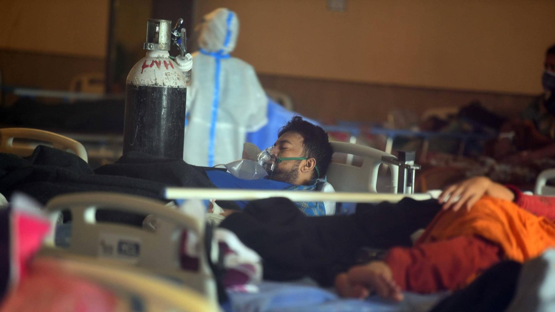 Ein Patient liegt in Indien mit Beatmungsmaske in einem Krankenzimmer, am Fußende eine große Sauerstoffflasche, im Hintergrund eine Person in Schutzanzug Ein Patient liegt in Indien mit Beatmungsmaske in einem Krankenzimmer, am Fußende eine große Sauerstoffflasche, im Hintergrund eine Person in Schutzanzug