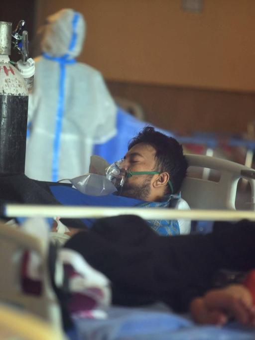 Ein Patient liegt in Indien mit Beatmungsmaske in einem Krankenzimmer, am Fußende eine große Sauerstoffflasche, im Hintergrund eine Person in Schutzanzug