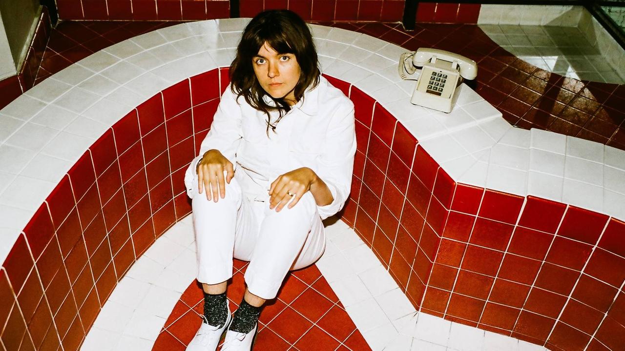 Courtney Barnett sitzt weiß gekleidet in einer rot weiß gekachelten Eckes eines Schwimmbades neben sich ein altes analoges Telephon Courtney Barnett sitzt weiß gekleidet in einer rot weiß gekachelten Eckes eines Schwimmbades neben sich ein altes analoges Telephon